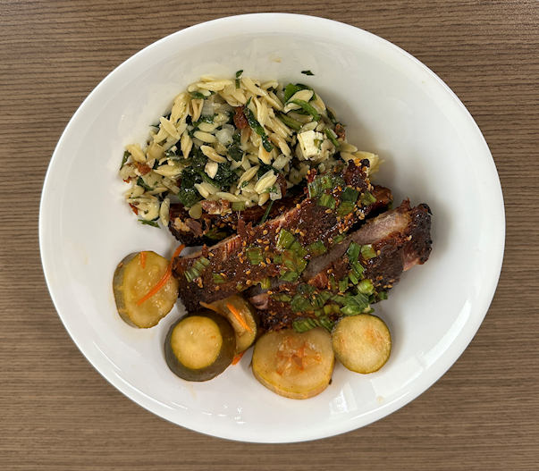 A white bowl containing three ribs, orzo salad, and spicy Korean Pickles.